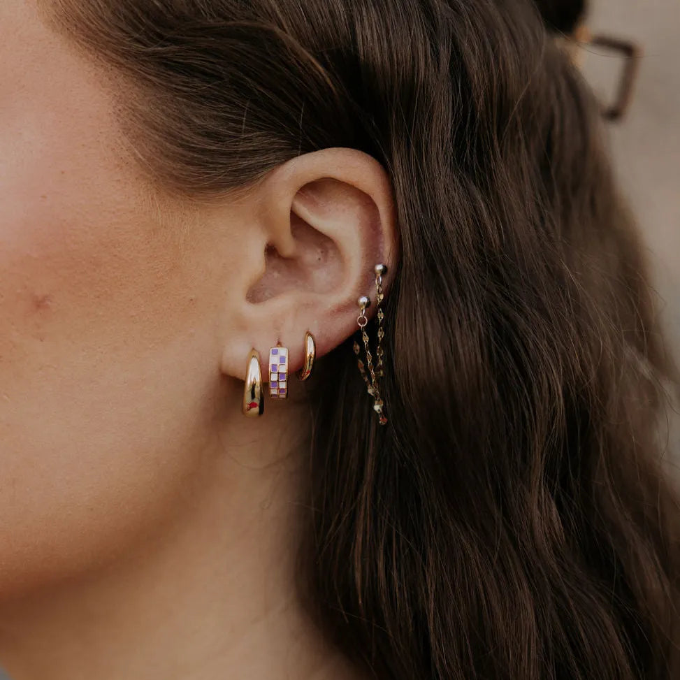 Close-up of an ear with multiple earrings including a pair of purple checkered hoops  and gold hoops against a blurred background