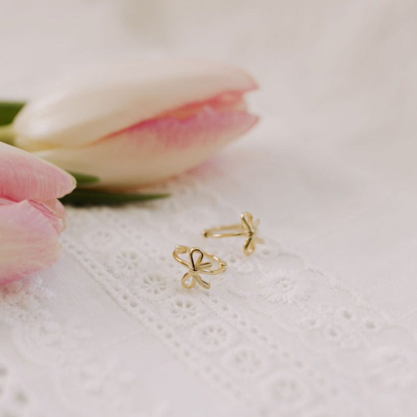 A pair of gold-colored hoop earrings with a bow detail, displayed on a white textured surface next to a pink tulip.