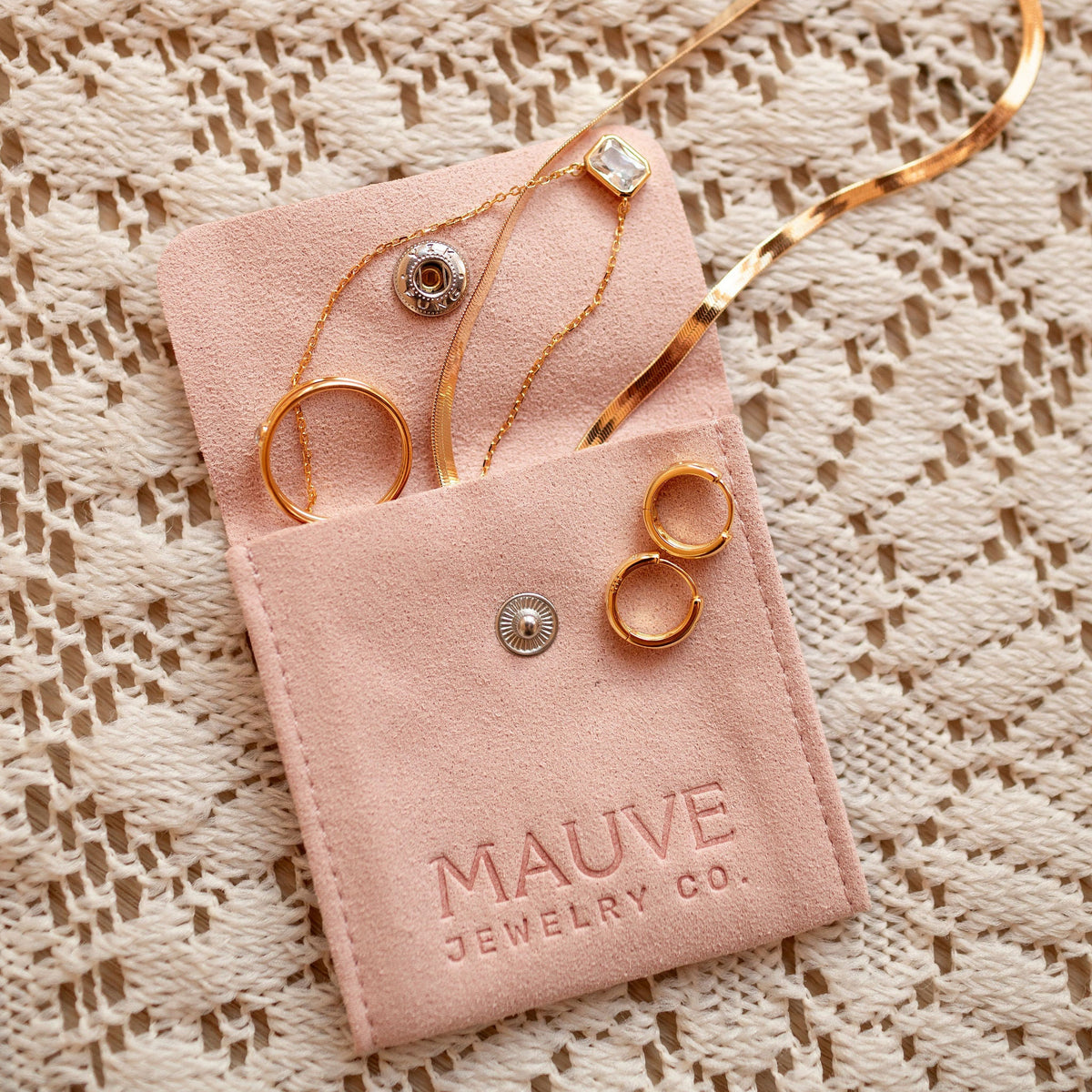 Gold earrings in a pink 'MAUVE JEWELRY CO.' pouch on a textured beige background