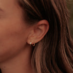 A close-up side profile of a woman with brown hair wearing a small gold hoop earring with a clear gemstone and a delicate gold chain necklace.