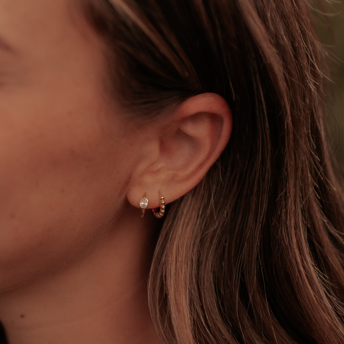 A close-up side profile of a woman with brown hair wearing a small gold hoop earring with a clear gemstone and a delicate gold chain necklace.