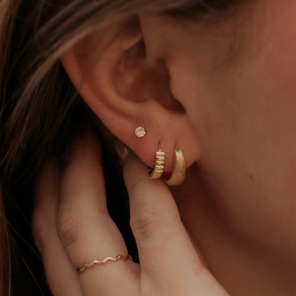 Close-up of an ear with gold hoop earrings and a ring, against a warm-toned background.