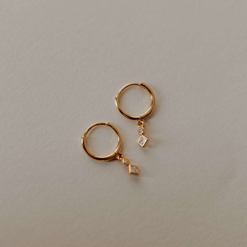Gold hoop earrings with small diamond-shaped charms on a light gray background