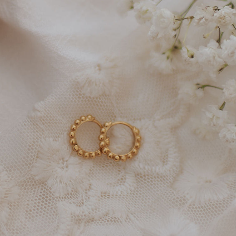 A pair of gold hoop earrings with a beaded textured border, displayed on a soft white lace fabric with white baby's breath flowers.