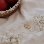 Two gold rings with textured, beaded edges resting on a white lace fabric, styled with white baby's breath flowers and a basket of red peaches in the background.