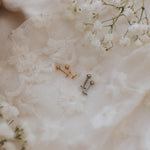 Two small barbell stud earrings, one gold-toned and one silver-toned, displayed on textured white lace fabric with white baby's breath flowers.