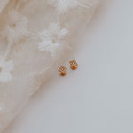 A pair of small square gold stud earrings with clear stones, resting on a white surface next to a cream-colored embroidered lace fabric.