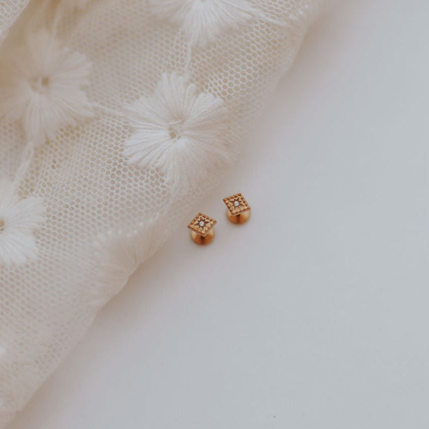 A pair of small square gold stud earrings with clear stones, resting on a white surface next to a cream-colored embroidered lace fabric.