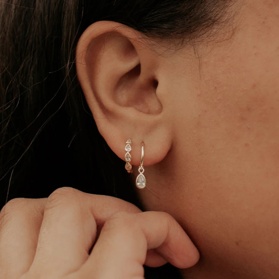 Close-up of an ear wearing gold hoop earrings with a teardrop-shaped pendant.