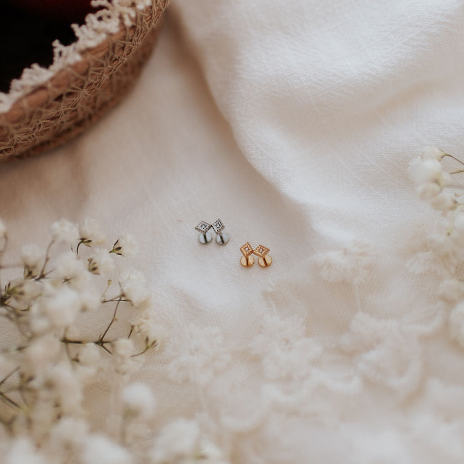 Two small stud earrings, one silver-toned and one gold-toned, displayed on a white lace fabric with baby's breath flowers and a woven basket of red fruit in the background.