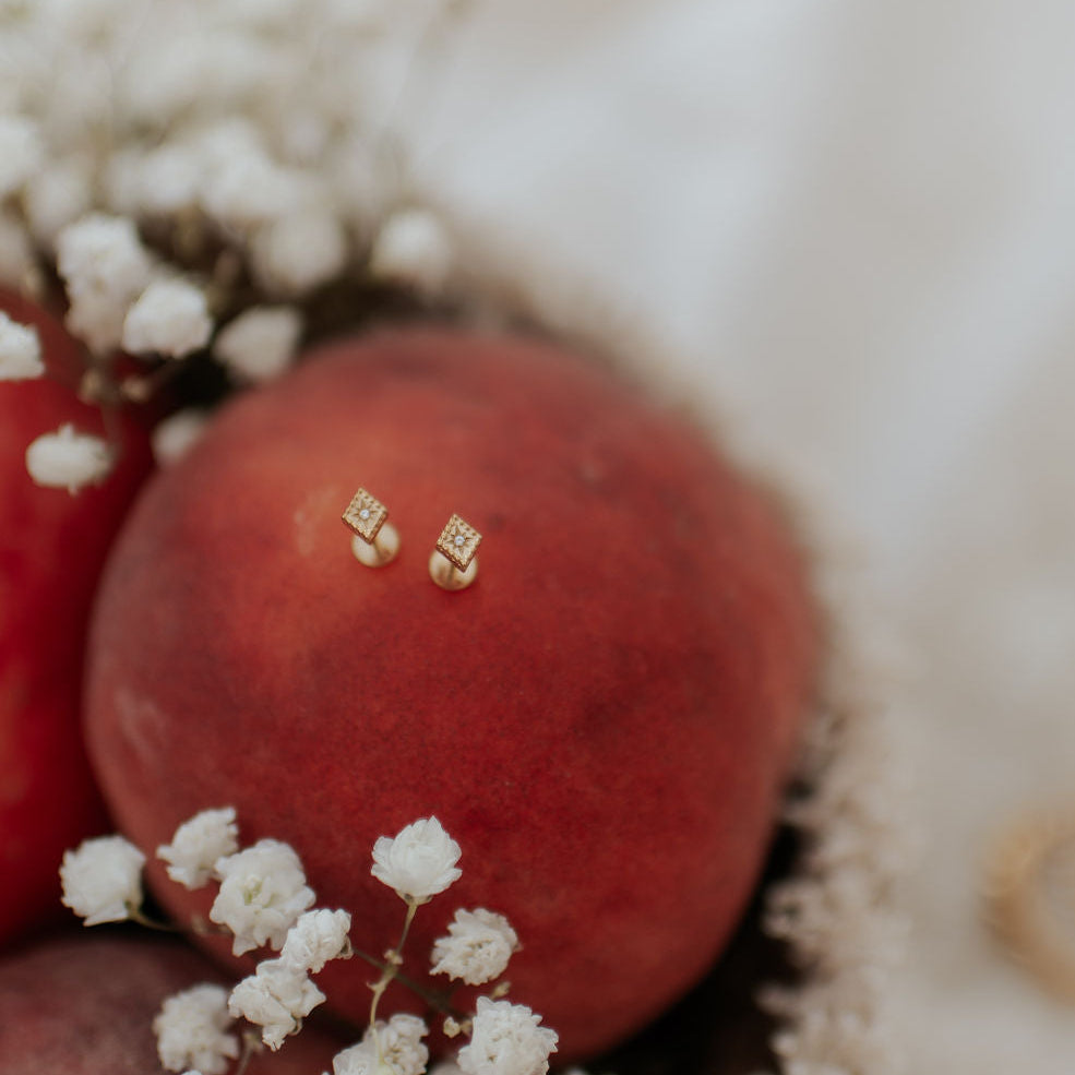A close-up of a pair of small gold stud earrings resting on the surface of a red fruit, surrounded by white baby's breath flowers on a soft white fabric background.
