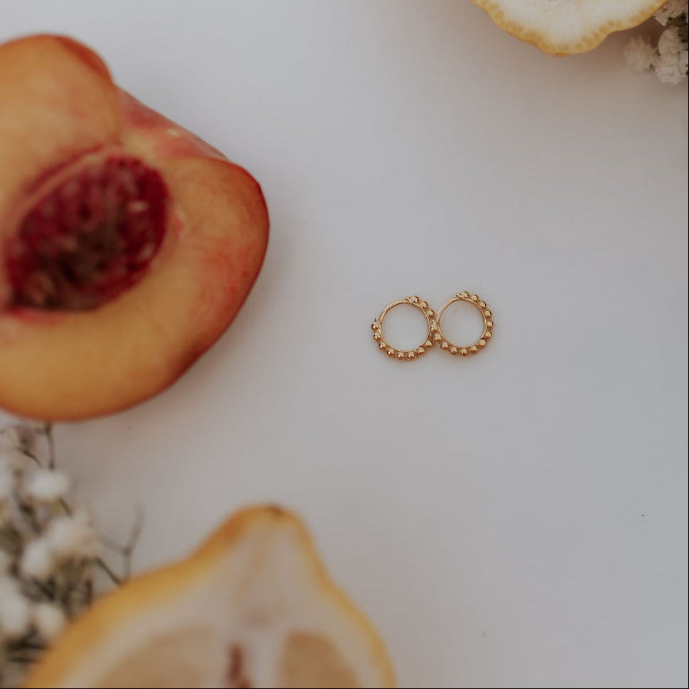 A pair of small gold hoop earrings with textured beaded edges, displayed on a clean white surface with decorative flat-lay props including a peach, lemon slices, and dried flowers.