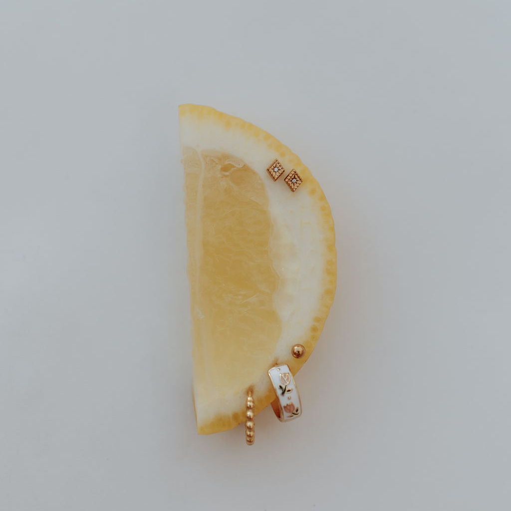 A pair of gold-toned stud earrings with floral enamel details, displayed on a yellow lemon wedge against a light gray background.