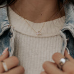 Person wearing a necklace with a small pendant, holding a phone, and wearing rings.