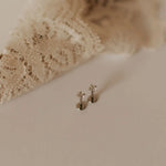 Two small silver flat back stud earrings on a textured beige surface with lace fabric in the background.