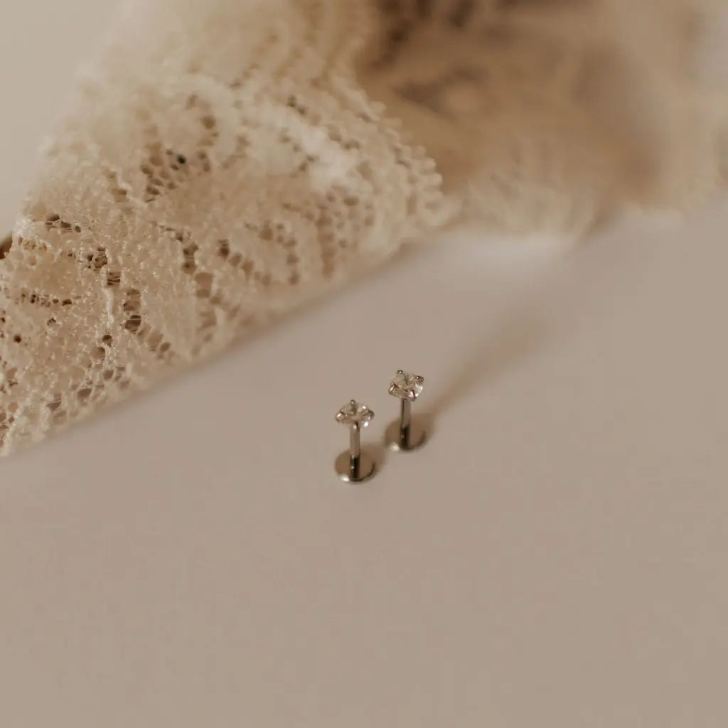 Two small silver flat back stud earrings on a textured beige surface with lace fabric in the background.