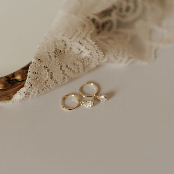 A pair of gold-colored hoop earrings on a white surface, with a hint of a lace curtain in the background.
