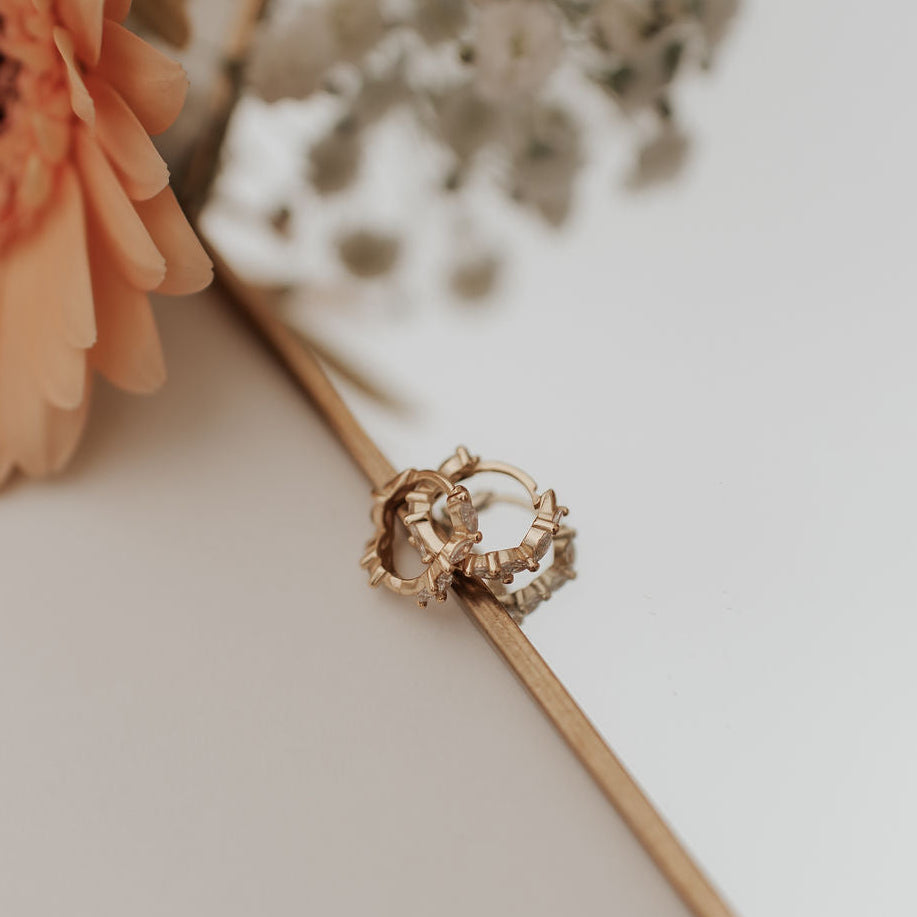 Gold diamond earrings with intricate design on a white surface with flowers in the background