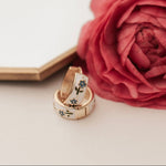 Two gold tea cup inspired earrings with gemstones on a white surface next to a pink rose.