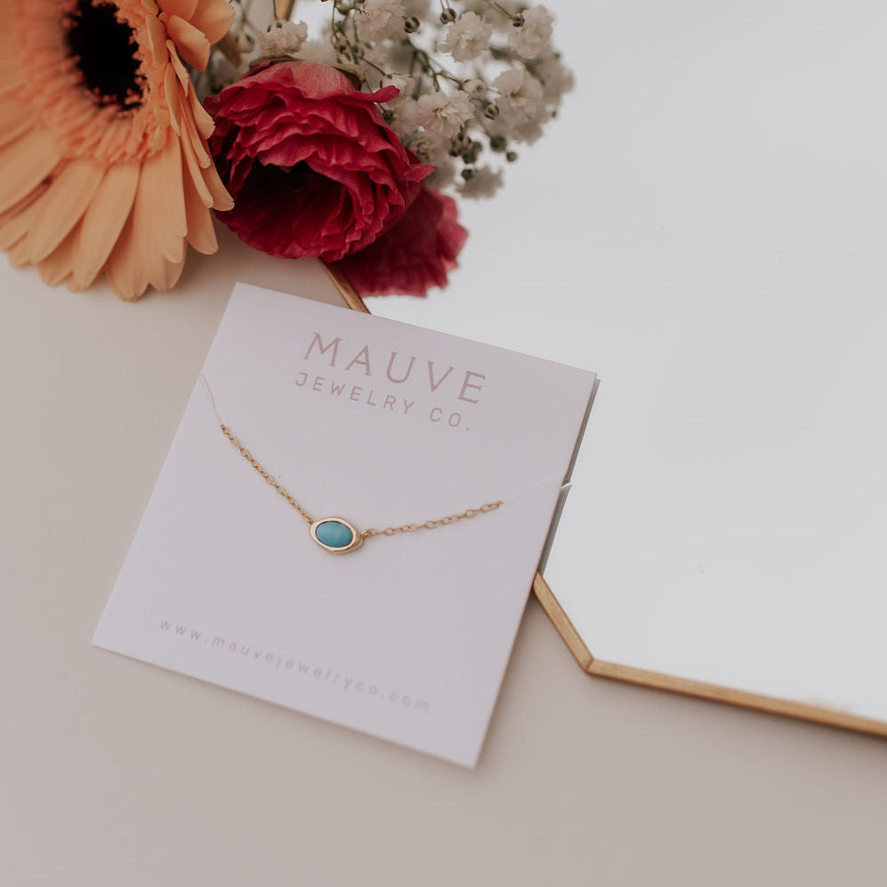 Gold necklace with a blue stone on a Mauve Jewelry Co. card with flowers in the background