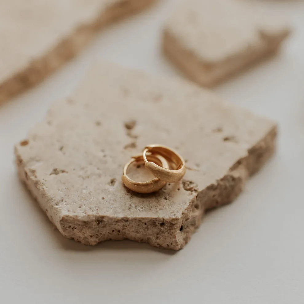 Two gold earrings on a textured stone surface with a blurred background