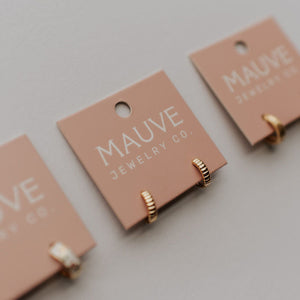 Three pairs of gold earrings on pink 'MAUVE JEWELRY CO.' cards against a light gray background.