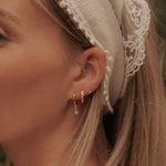 Close-up of a woman's ear wearing small gold hoop earrings, one with a dangling geometric charm and another with a textured vertical bar, styled with a cream lace headband.