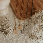 Three gold necklaces with engraved pendants on a textured surface with dried flowers.