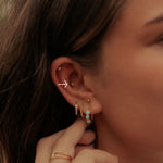 Close-up of a person's ear adorned with multiple delicate earrings, including rose gold hoop, stud, and drop earrings with light blue stones, while a hand gently touches the ear.