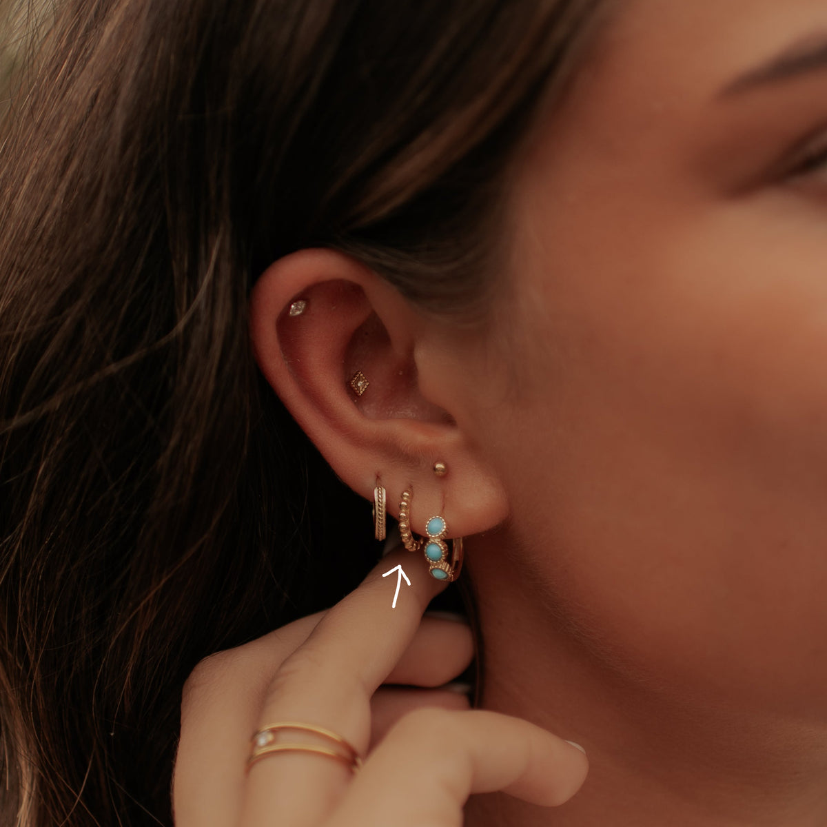 A close-up of a woman's ear adorned with multiple gold piercings, including a small hoop, a huggie, and a cluster of turquoise and gold beads, with her hand resting near her neck.