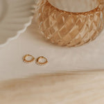 Gold pearl hoop earrings on a beige surface with a textured gold object in the background
