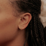 A close-up side profile of a person with dark braided hair wearing a delicate gold huggie hoop earring and a small gold stud, with a thin gold chain necklace visible at the neck.