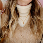 Close-up of a person wearing a beige turtleneck sweater and patterned cardigan with layered gold necklaces.