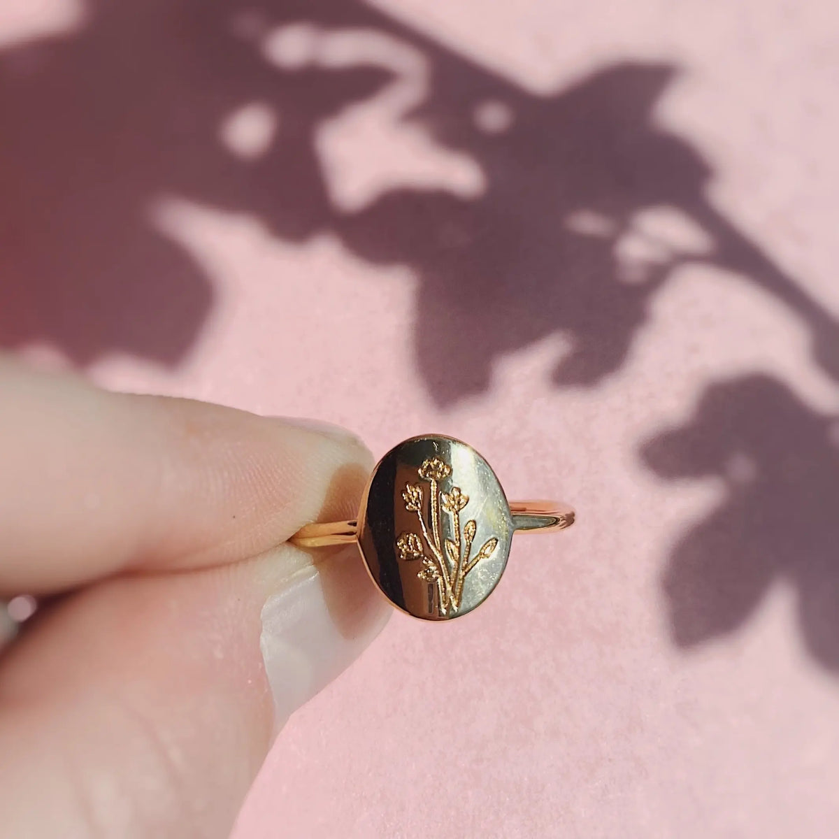 Gold ring with floral design held by a hand on a pink background