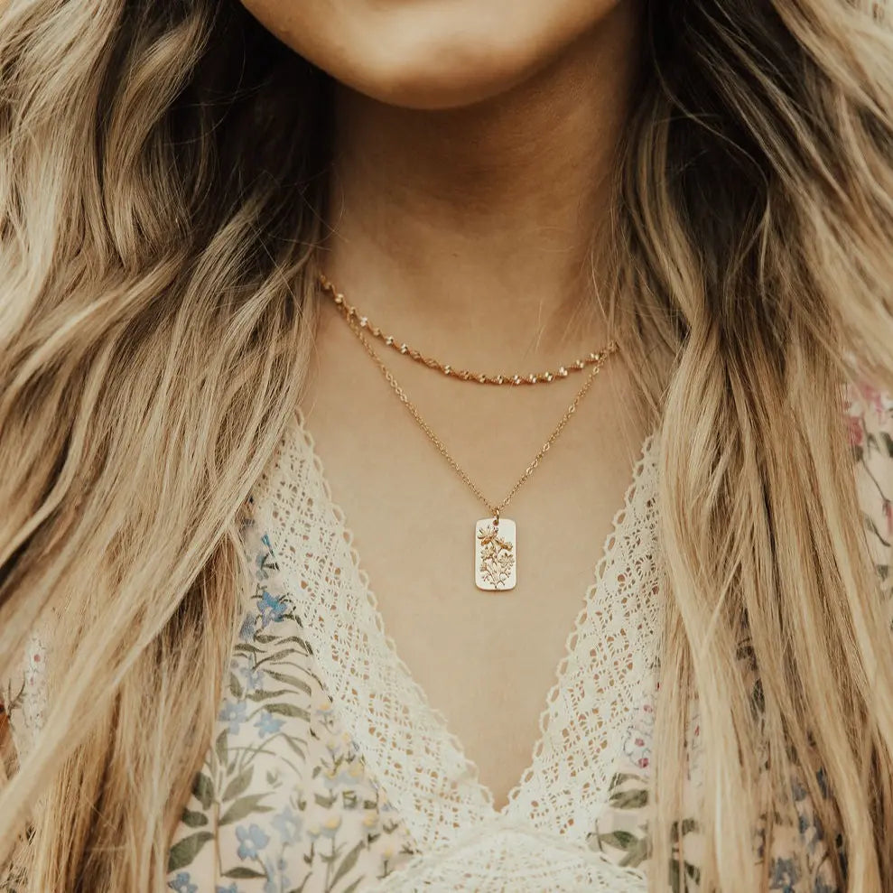 Close-up of a person wearing a gold necklace with a tag pendant, with long blonde hair and a floral top.