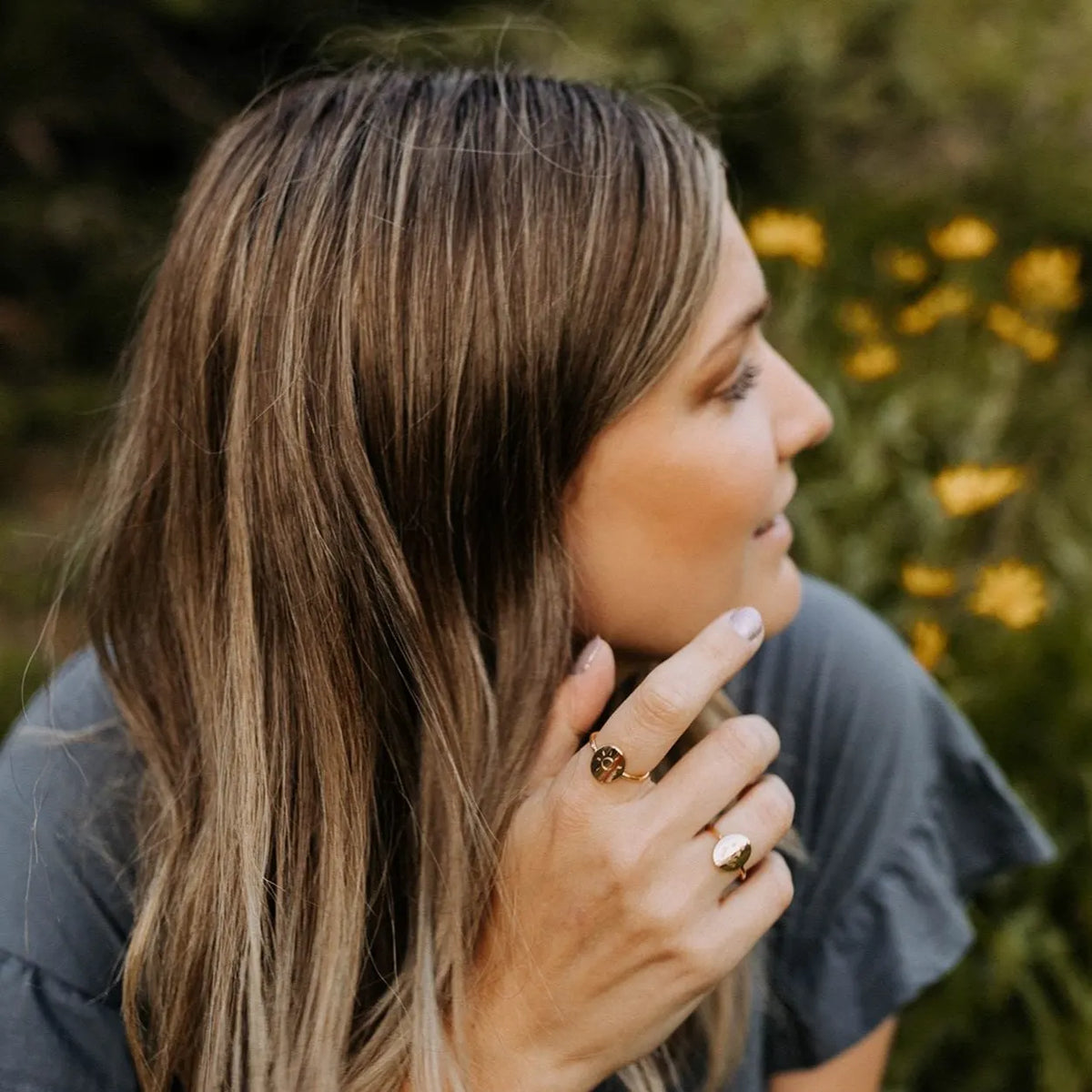 Woman with long hair and rings, sitting outdoors with blurred flowers in the background