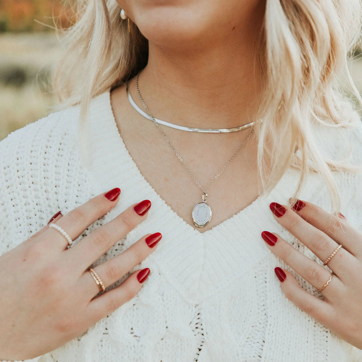 Woman wearing a white sweater with gold necklaces and rings, outdoors.