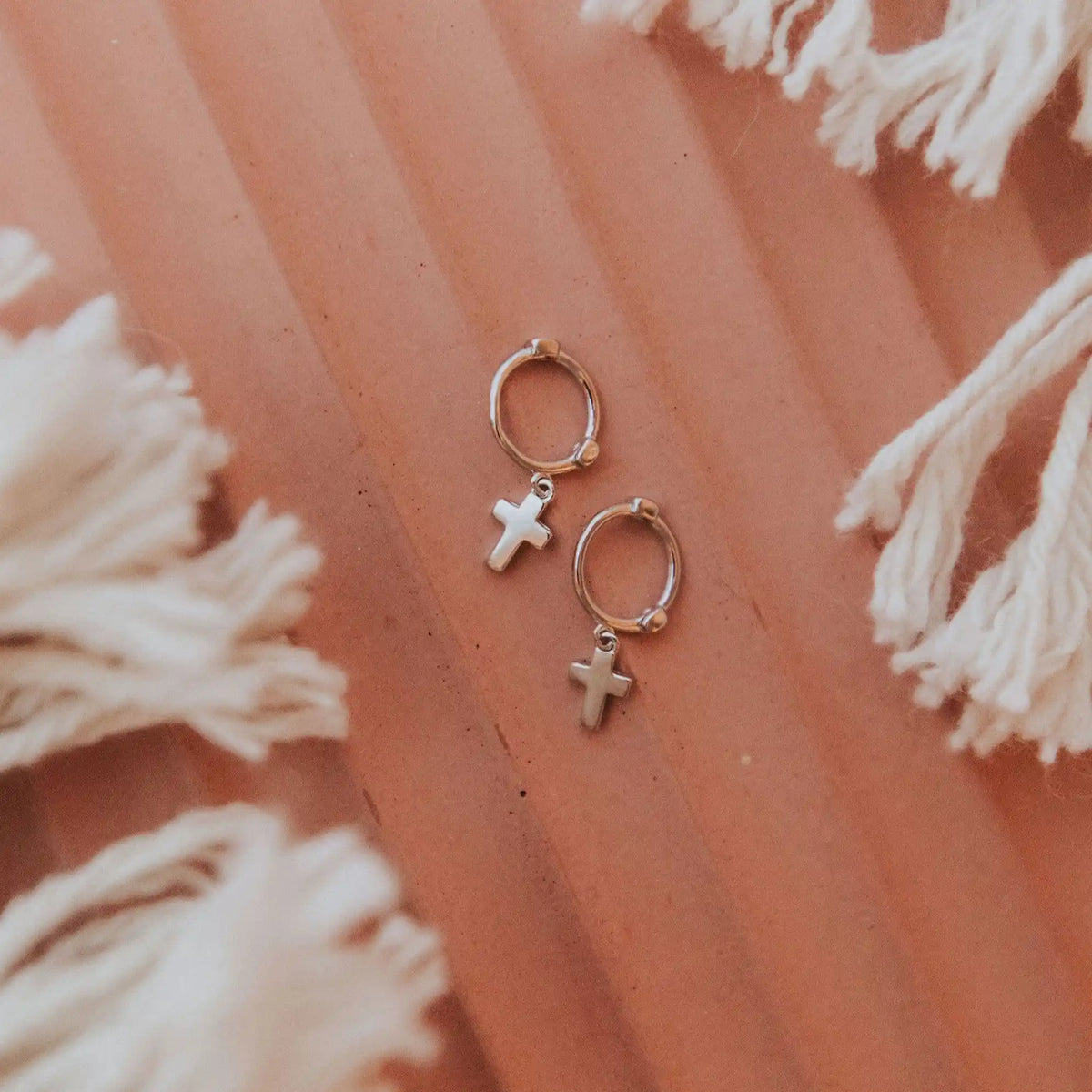 Small hoop earrings with cross charms on a textured pink surface with white tassels.