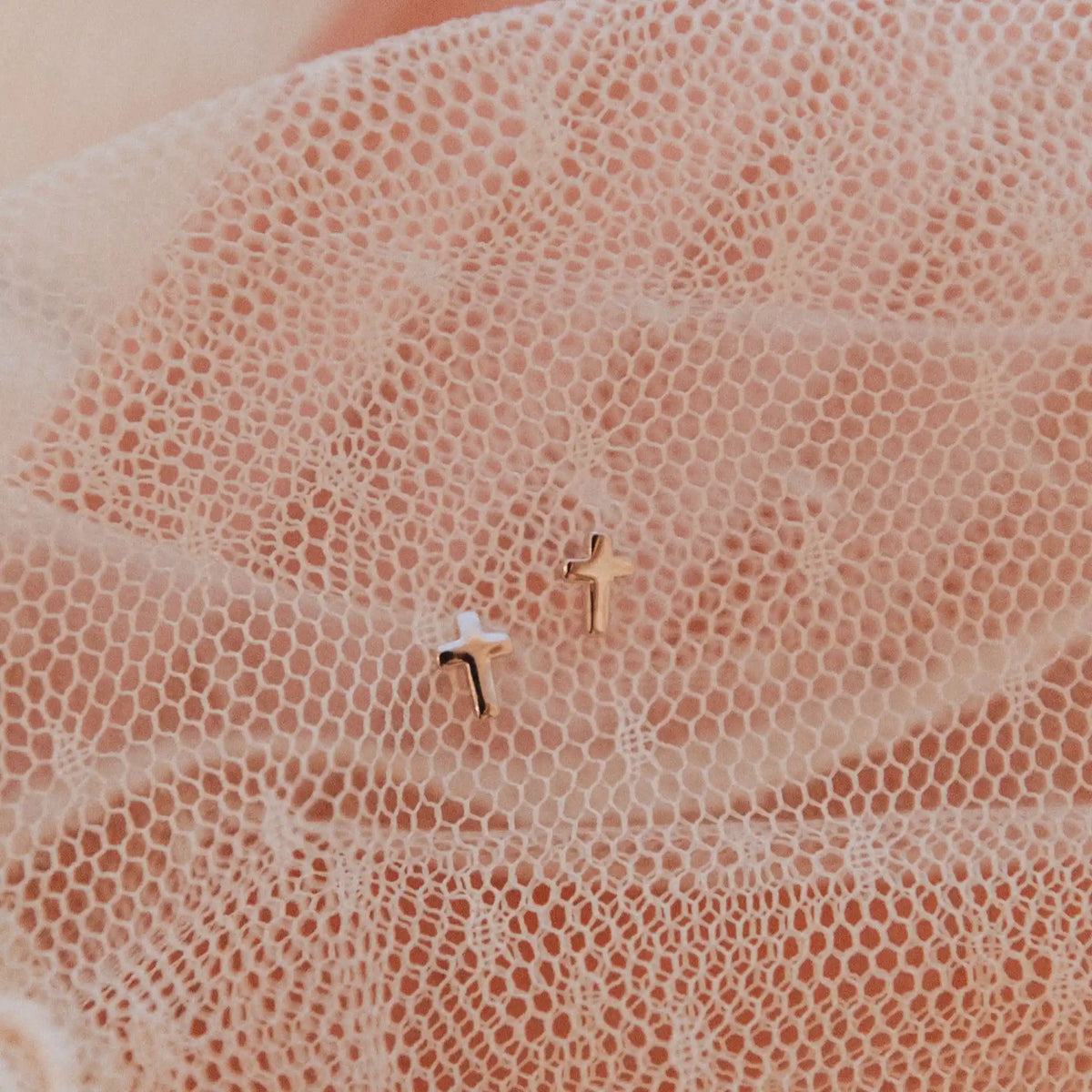 Two small gold cross earrings on a textured beige surface