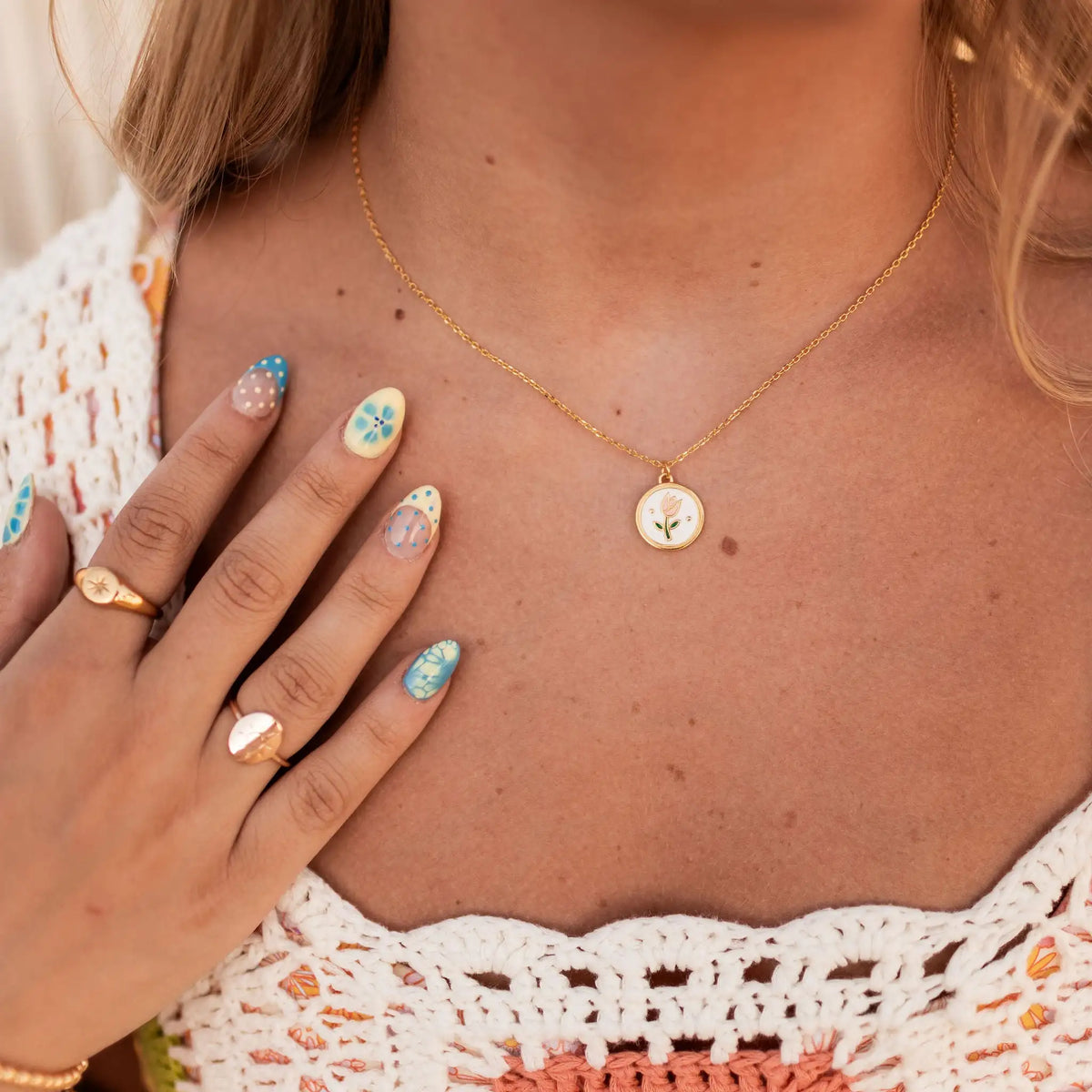 Close-up of a person wearing a gold necklace with a small pendant, hand with colorful nail polish, and rings.