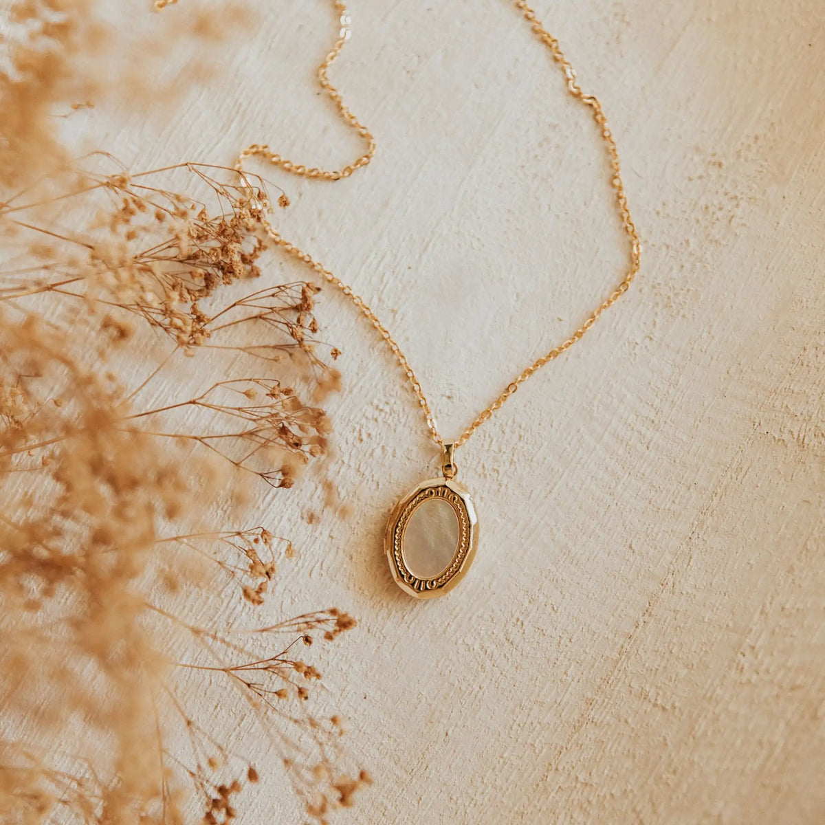 Gold necklace with a pendant on a textured beige surface with dried flowers.
