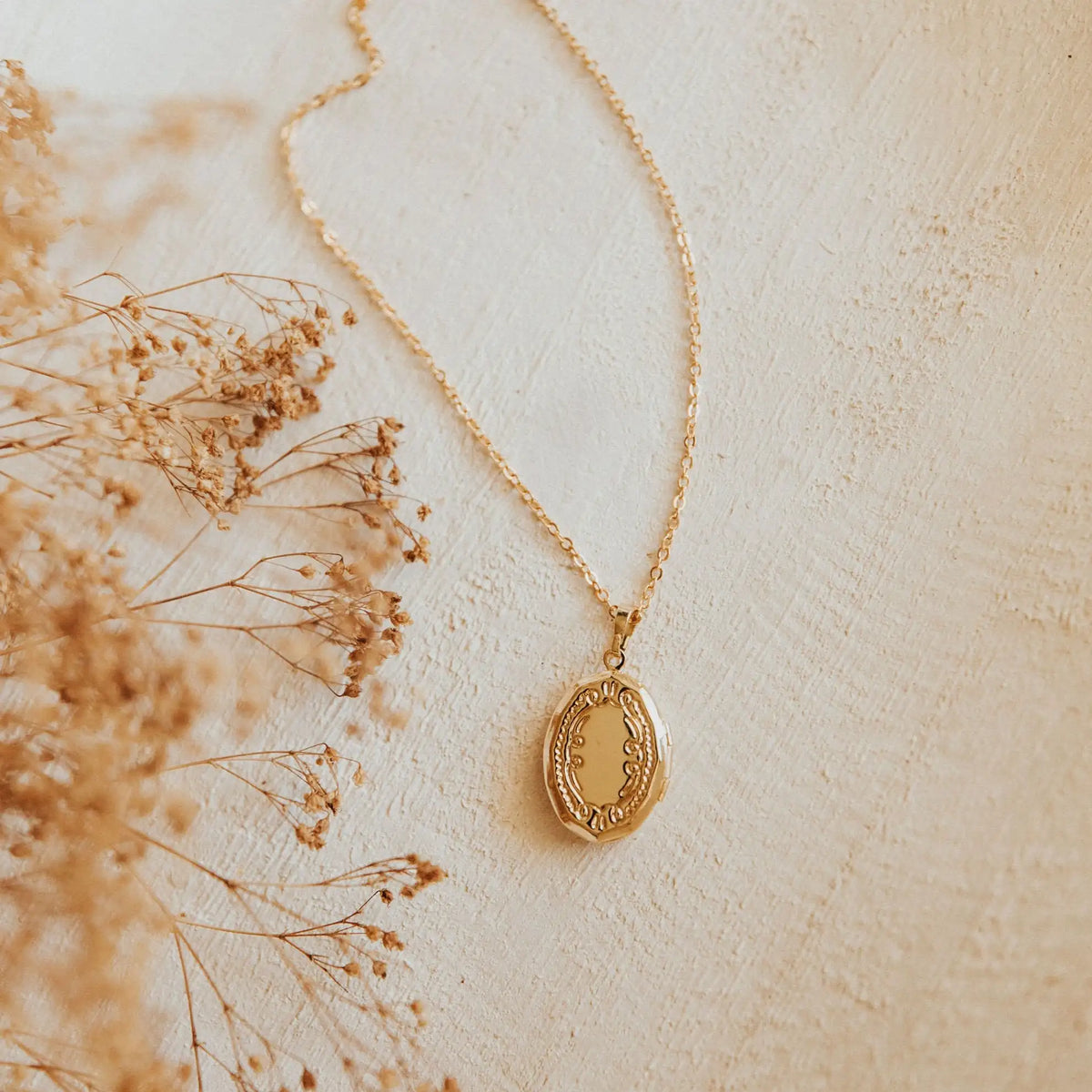 Gold necklace with a pendant on a light beige background with dried flowers.