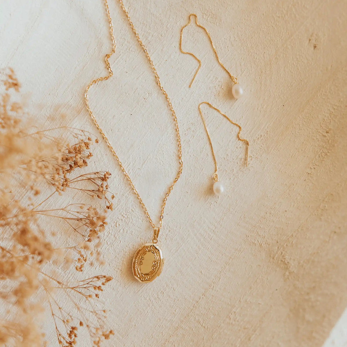 Gold necklace with a pendant on a textured beige surface with dried flowers.