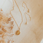 Gold necklace with a pendant on a textured beige surface with dried flowers.