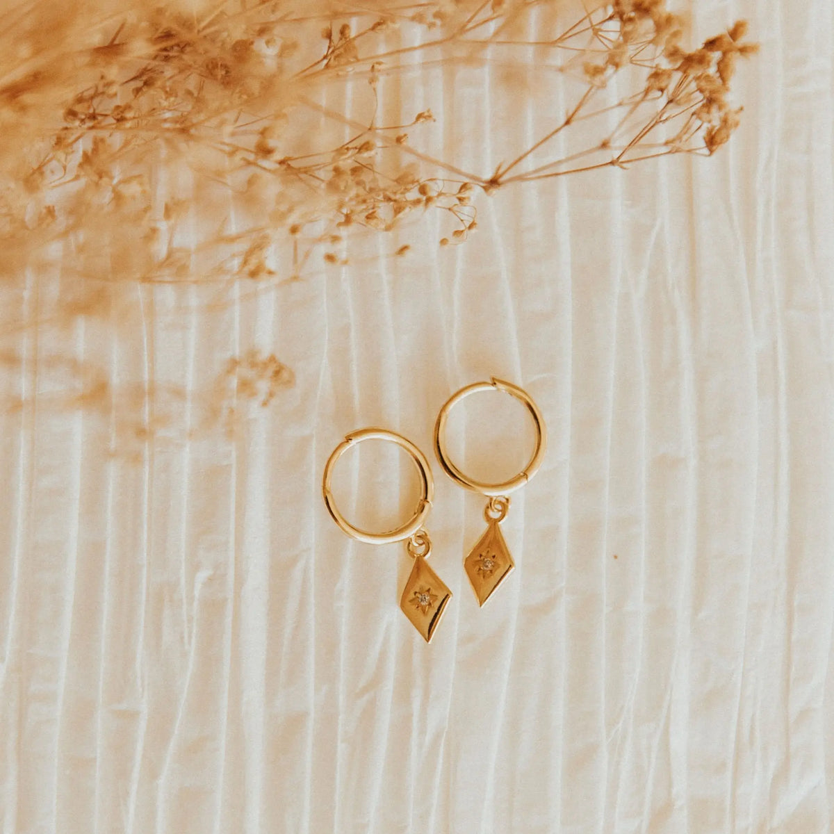 Gold hoop earrings with diamond-shaped charms on a beige background