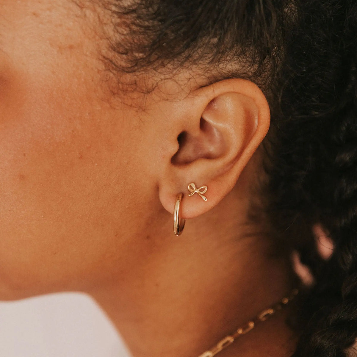 Close-up of an ear wearing gold hoop earrings with a bow detail.