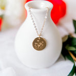 Gold necklace with a circular pendant on a white vase against a blurred floral background