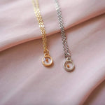 Two necklaces, one gold and one silver, on a soft pink fabric background.