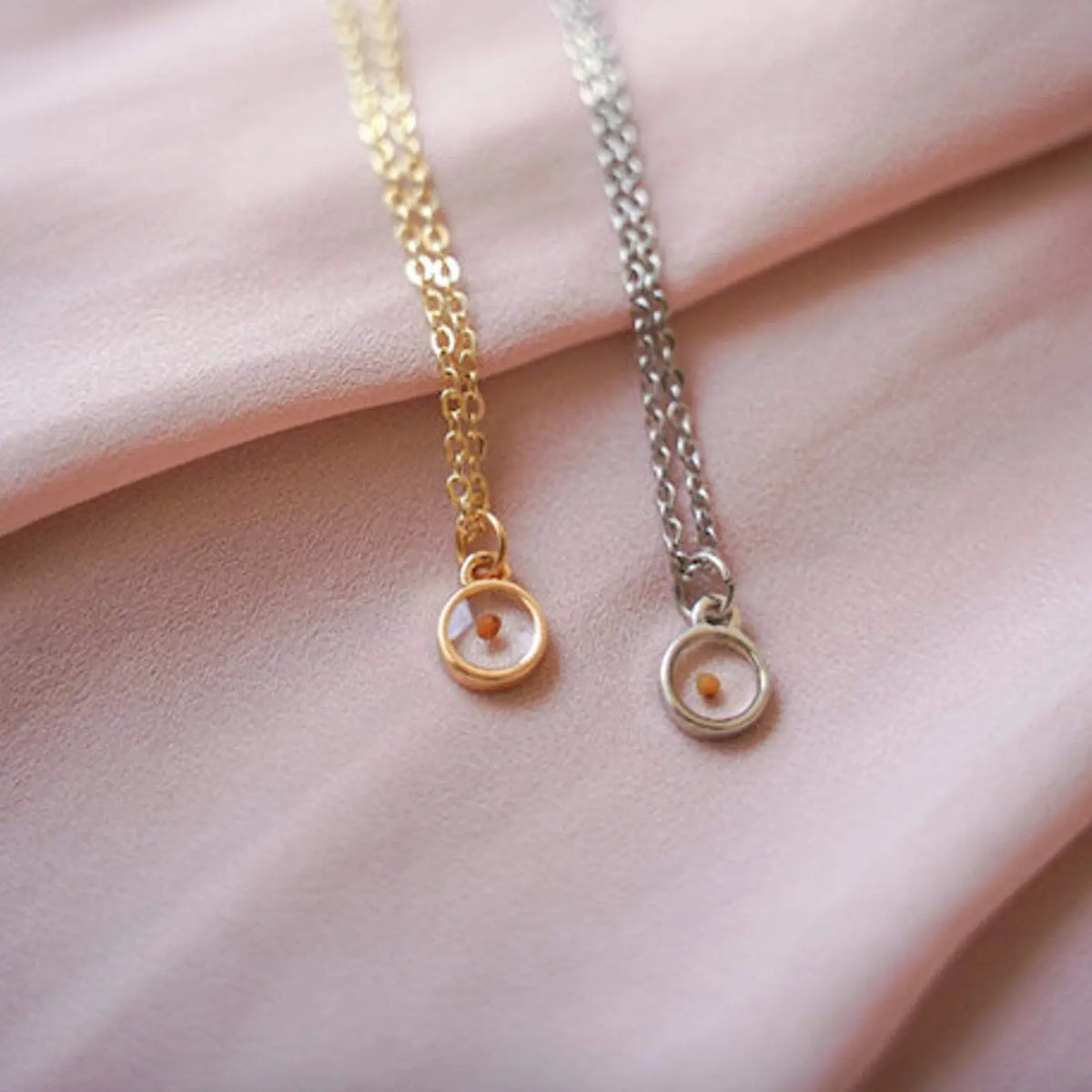 Two necklaces, one gold and one silver, on a soft pink fabric background.