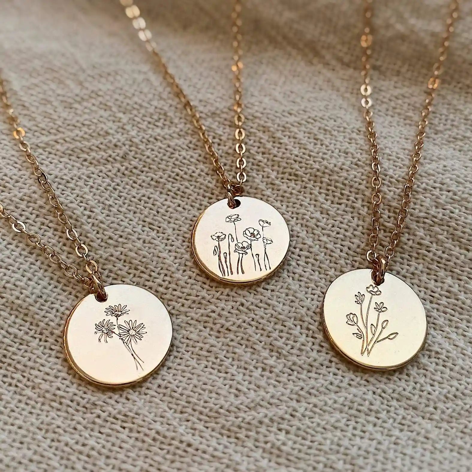Three gold necklaces with circular pendants featuring floral designs on a textured beige fabric background.